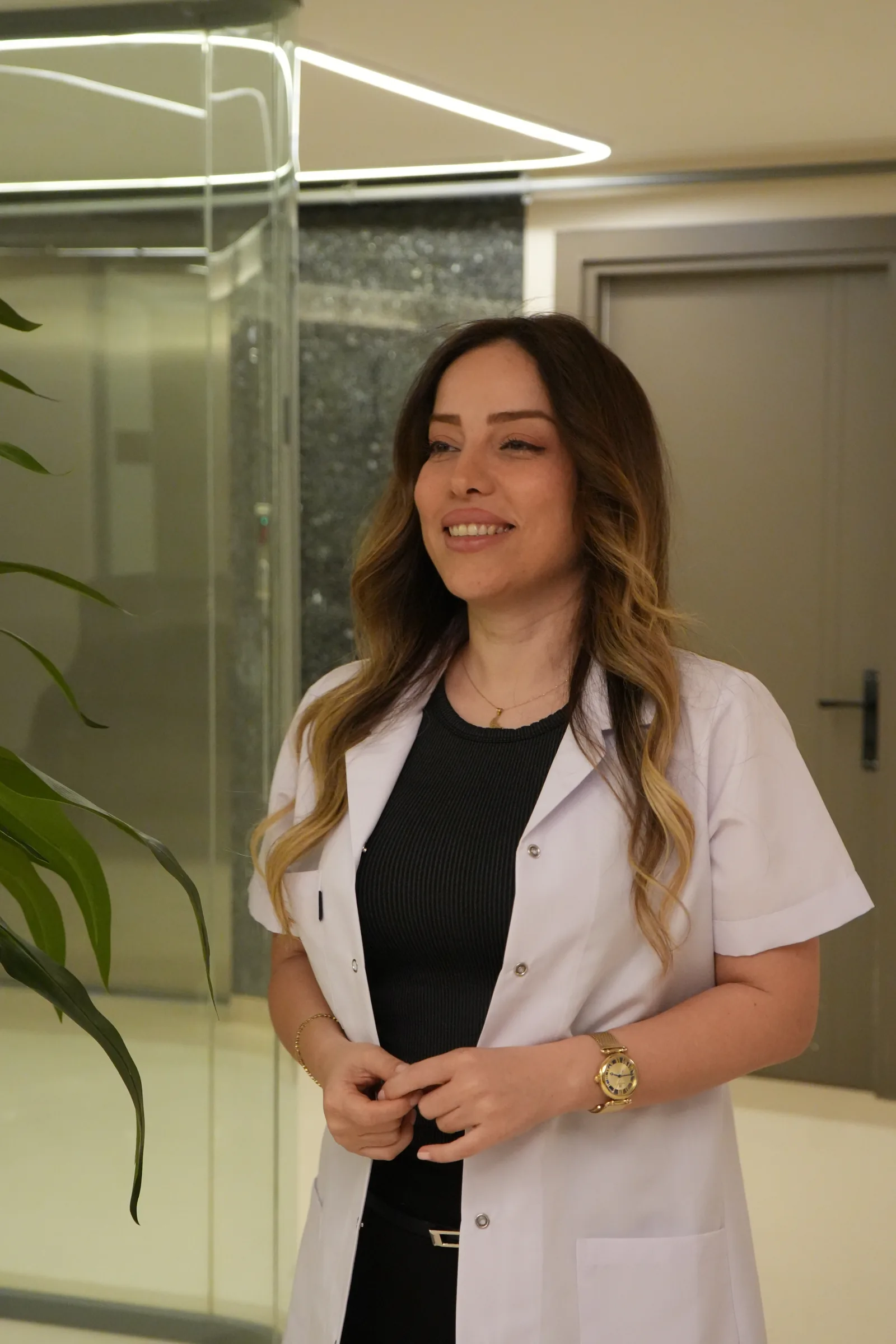 Azin Asgari klinikte, beyaz önlüğüyle, gülümseyerek yana bakıyor.