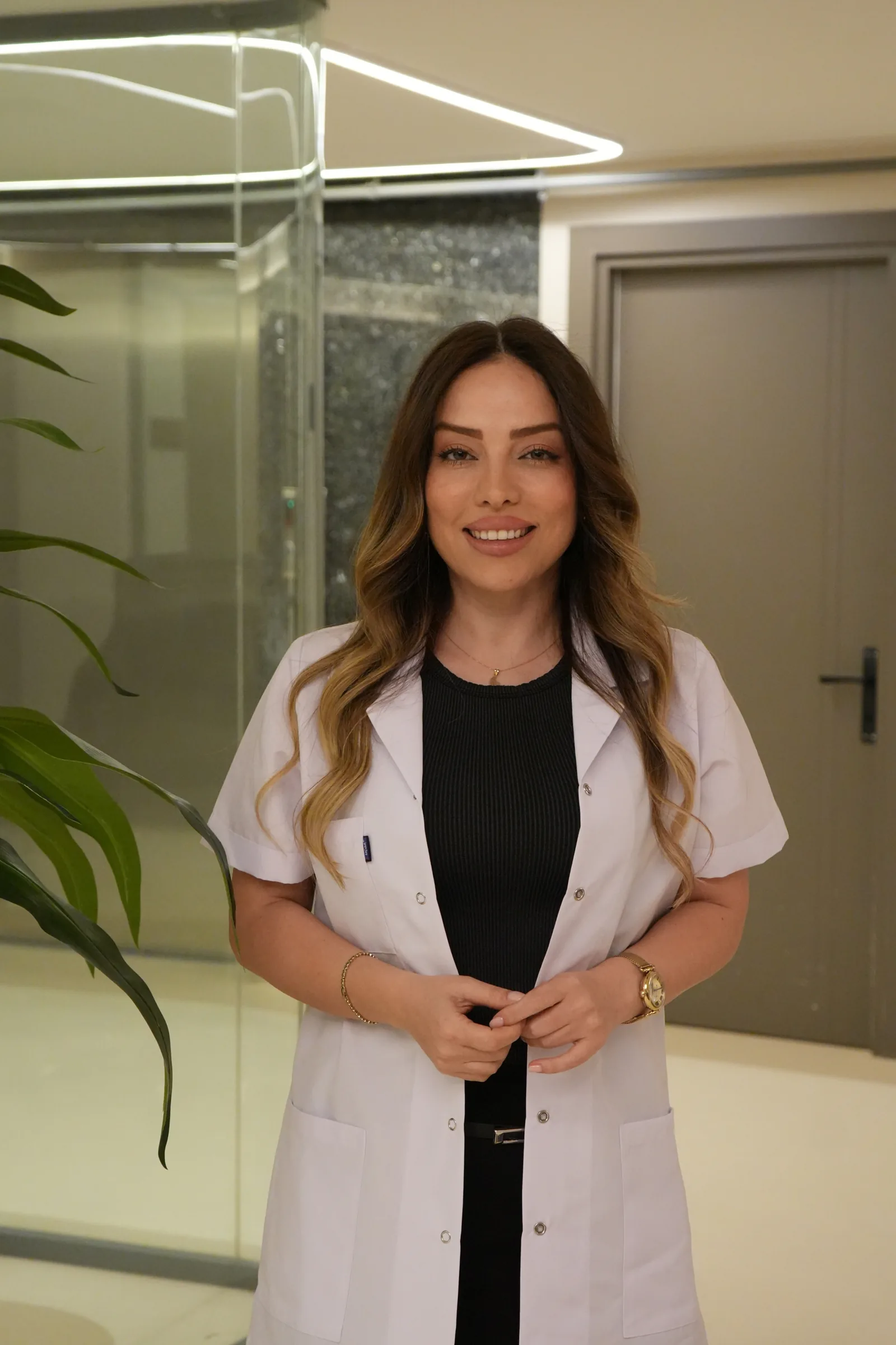 Azin Asgari, beyaz önlüğüyle kliniğin koridorunda, kameraya bakarak gülümsüyor.
